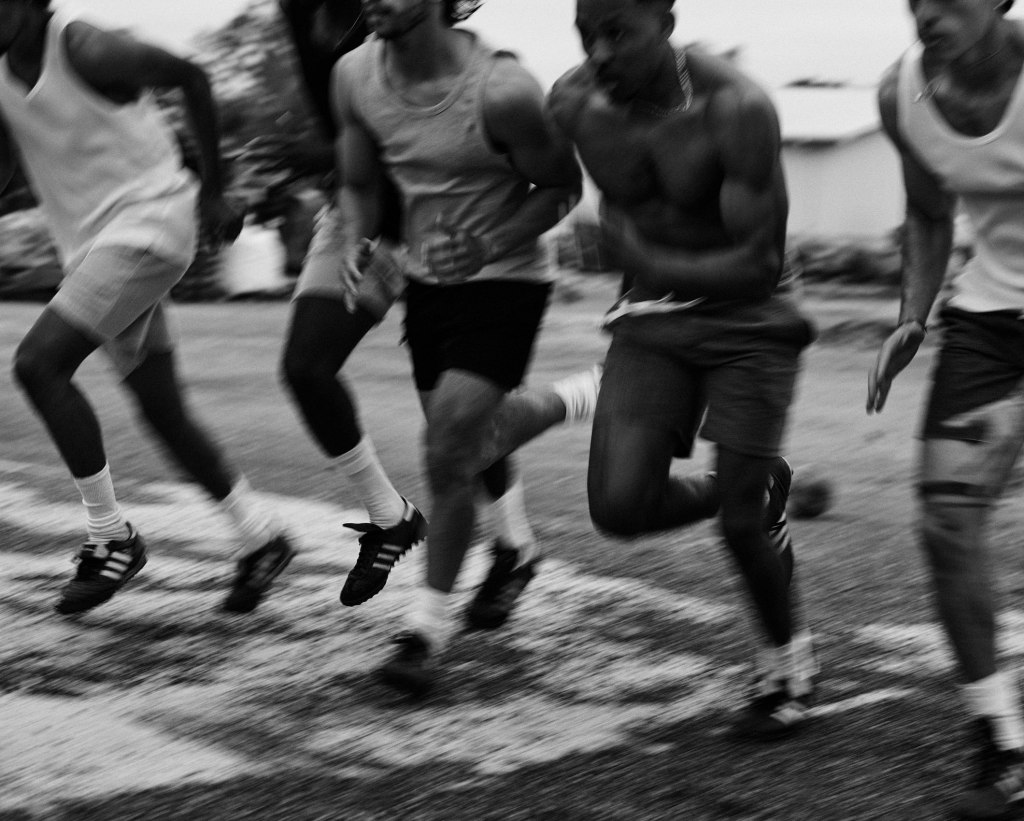 A black-and-white photo capturing a dynamic moment as five young men sprint forward from a starting line. The motion blur emphasizes speed and energy. They wear athletic clothing and running shoes, with a mix of tank tops, shorts, and one runner shirtless. The background is slightly out of focus, showing a street or outdoor area with some buildings and vegetation.
