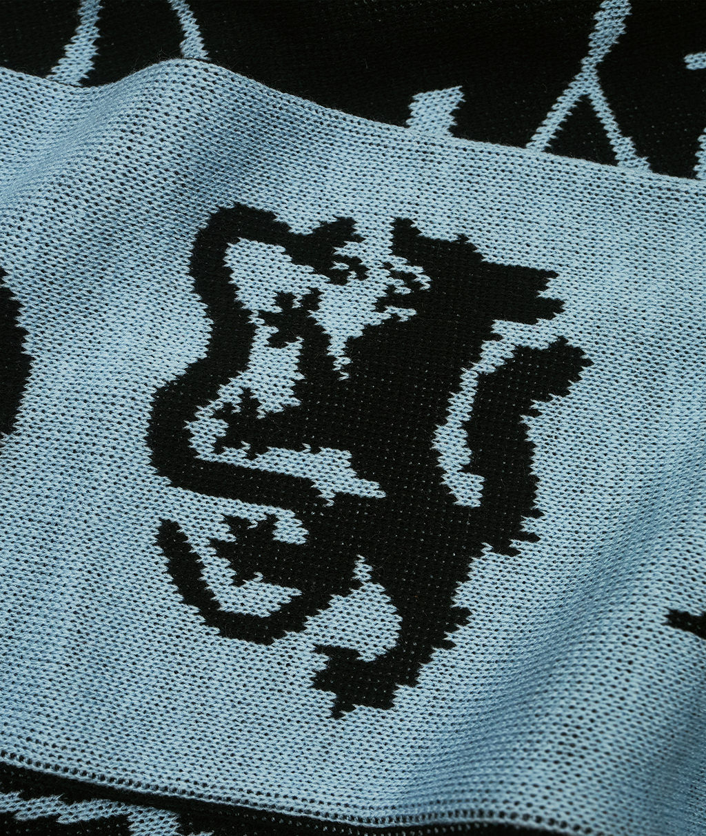 A close-up image of a knitted fabric featuring a black and light blue design. The pattern includes a stylized heraldic lion or wolf-like figure, woven in black against the light blue background. The texture of the knit and the slight fold in the fabric are clearly visible, adding depth and movement to the image.