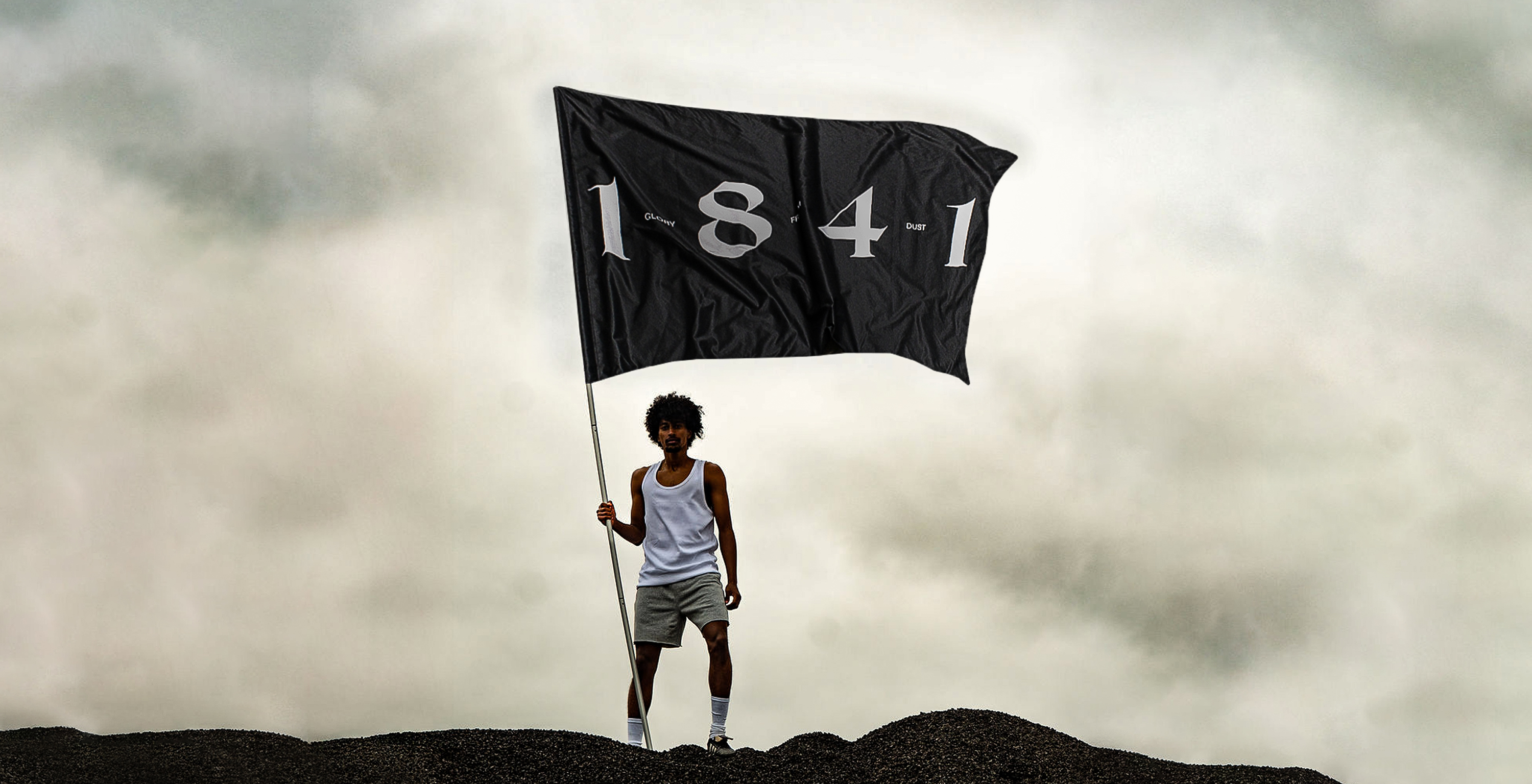 A person stands on a dark, rocky hill under an overcast sky, holding a large black flag that reads "1841" in bold silver letters. Smaller text between the numbers reads "GLORY," "FROM," and "DUST." The individual wears a white tank top, gray shorts, and white socks with sneakers.
