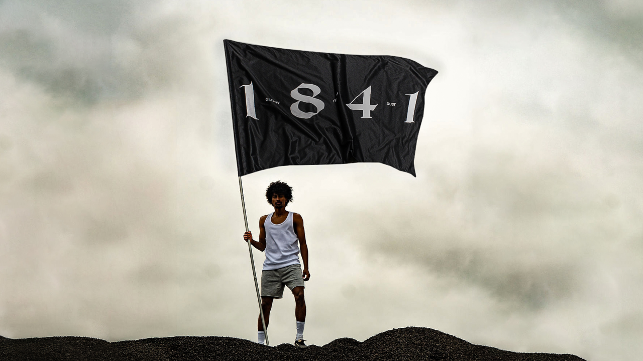 A man in a white tank top and gray shorts stands on a rocky hill holding a large black flag with the number “1841” printed in bold white letters. The flag waves against a cloudy sky backdrop.